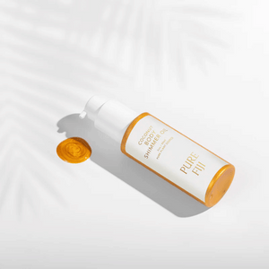 Bottle of Coconut Body Shimmer Oil | Pure Fiji with golden shimmer oil drop on a white surface with soft palm leaf shadows, showcasing skincare and body care product.