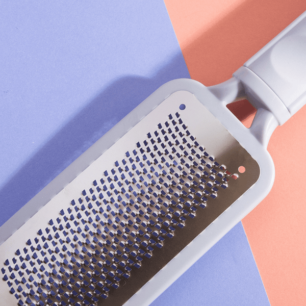 Close-up of the stainless steel grating surface and white handle of The Gratest Professional Micro-Grated XL Rasp | Barefoot Scientist against a pastel blue and peach background.