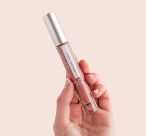 Hand holding a tube of Vitamin Glaze® Oil Infused Lip Gloss – Delicate Rose | Farmhouse Fresh with a soft pink background, highlighting its delicate rose shade and sleek design for skincare and beauty.