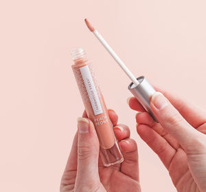 Hands holding a tube and applicator of Vitamin Glaze® Oil Infused Lip Gloss – Peach Peony | Farmhouse Fresh with peachy nude gloss on a soft pink background