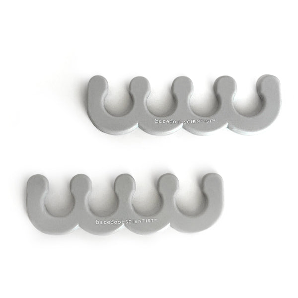 Two gray silicone toe separators designed for pedicure use, each featuring a curved, wavy shape with the text "barefoot SCIENTIST" printed on them, part of the Spaced Out | Barefoot Scientist collection.