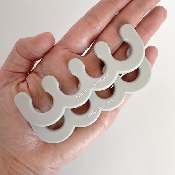 A close-up of a hand holding two gray, curved silicone toe separators branded with "barefoot scientist," designed for foot wellness and recovery, Spaced Out | Barefoot Scientist.