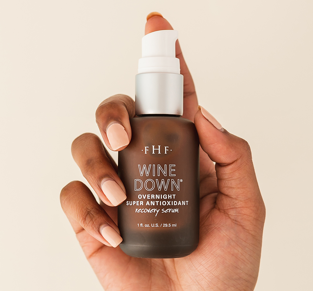 Hand holding a frosted brown glass bottle with a white pump dispenser labeled Wine Down® Overnight Super Antioxidant Recovery Serum | Farmhouse Fresh, featuring nude-colored nails.