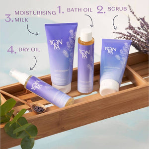 Set of four lavender-scented skincare products on a wooden bath tray including Phyto Bain - Invigorating, Relaxing Aromatic Bath I Yon-Ka Paris bath oil, scrub, moisturizing milk, and dry oil with natural herbs in the background.