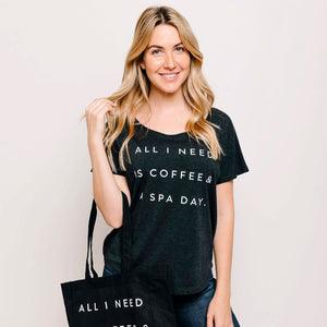 Woman wearing a black "Coffee & A Spa Day Women's T-Shirt | Lucky Owl" with white text that reads "All I need is coffee & a spa day," smiling and holding a black tote bag with matching text.