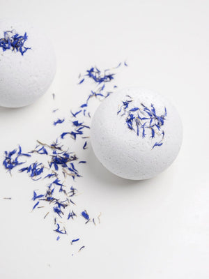Two white Snooze Bath Bomb | Bathorium with blue flower petals on top and scattered petals on a white surface, evoking a calm and soothing wellness atmosphere.
