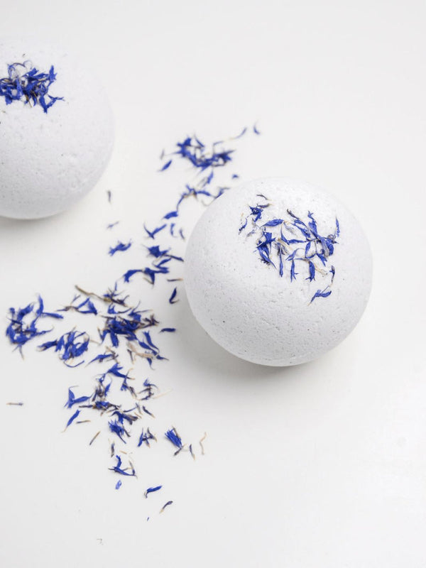 Two white Snooze Bath Bomb | Bathorium with blue flower petals on top and scattered petals on a white surface, evoking a calm and soothing wellness atmosphere.