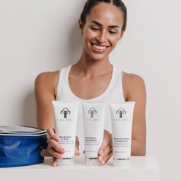 Smiling woman in white tank top holding three skincare products displayed on a white surface with a blue bag nearby, featuring the Body Regimen Bundle | Circadia.