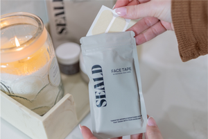 Hand holding a pair of light beige Face Tape Smoothing Strips | Seald™ Wellness above a silver package of the product, with a lit candle and skincare items blurred in the background.