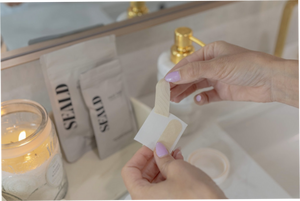 Person peeling a beige Face Tape Smoothing Strip | Seald™ Wellness from its backing in a bathroom setting with skincare products and a lit candle nearby.