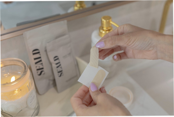 Person peeling a beige Face Tape Smoothing Strip | Seald™ Wellness from its backing in a bathroom setting with skincare products and a lit candle nearby.