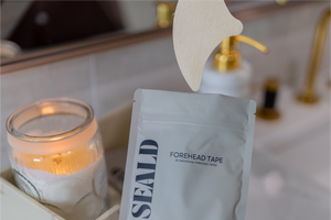 Package of Forehead Tape – Wrinkle Smoothing Patches | Seald™ Wellness displayed on a bathroom counter next to a lit candle and spa tool, promoting skincare and wellness.