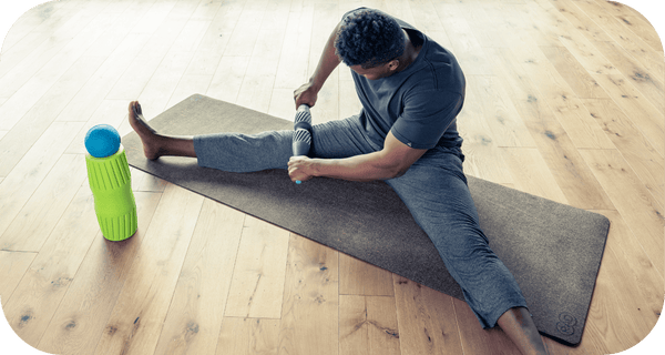 Man using a textured RAD HELIX | RAD massage roller on his leg while sitting on a yoga mat in a bright wooden floor room for muscle recovery and wellness.