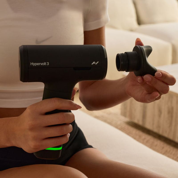 Person holding a black Hypervolt 3 | Hyperice percussion massage device with an attachment in a cozy indoor setting, promoting wellness and body care recovery.
