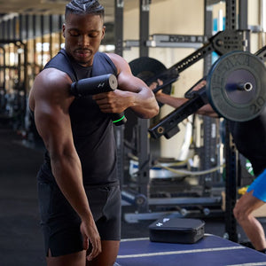 Athletic man using a Hypervolt 3 Pro | Hyperice massage device on his upper arm in a gym setting with weightlifting equipment in the background.