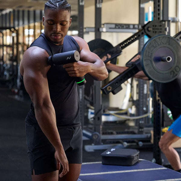 Athletic man using a Hypervolt 3 Pro | Hyperice massage device on his upper arm in a gym setting with weightlifting equipment in the background.