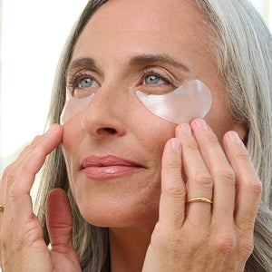 Mature woman applying FLASHPATCH® REJUVENATING EYE GELS | Patchology under her eyes, showcasing skincare treatment for eye rejuvenation and depuffing.