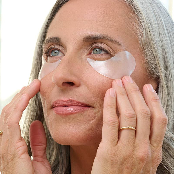 Mature woman applying FLASHPATCH® REJUVENATING EYE GELS | Patchology under her eyes, showcasing skincare treatment for eye rejuvenation and depuffing.