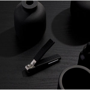 Precision Grip Toenail Clipper | Tweezerman with textured black handle for secure hold, placed on a matte black surface alongside minimalistic black decor items.