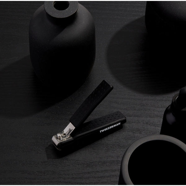 Precision Grip Toenail Clipper | Tweezerman with textured black handle for secure hold, placed on a matte black surface alongside minimalistic black decor items.