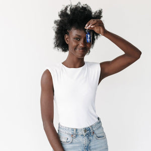 Smiling woman holding a small blue bottle of Vitamin C Reversal Serum (Professional) | Circadia near her forehead, wearing a white sleeveless top and light denim jeans.